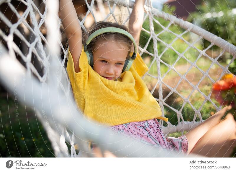 Junges Mädchen genießt Musik in einer Hängematte im Hinterhof Kind jung entspannend Kopfhörer Freizeit im Freien Pullover gelb Komfort gemütlich Genuss