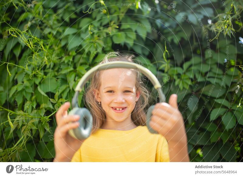 Lächelndes Mädchen mit Kopfhörern im Freien vor grüner Kulisse Daumen hoch Grün heiter jung Kleid gelb Beteiligung Garten Blatt Natur Sommer Fröhlichkeit Kind