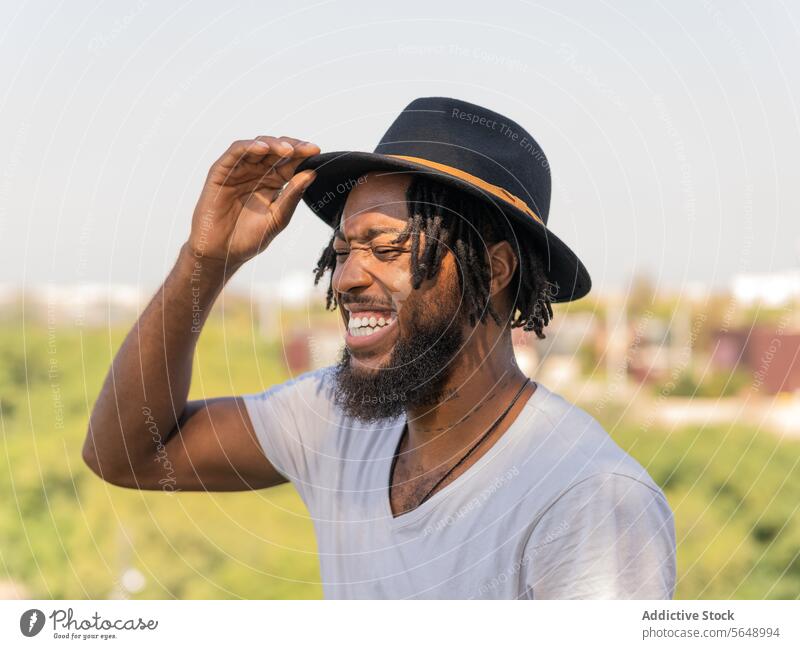 Fröhlicher schwarzer Mann, der seinen Hut im Park zurechtrückt ausrichten fixieren Sonnenlicht Lachen Spaß haben Witz Humor lustig berühren Lächeln positiv