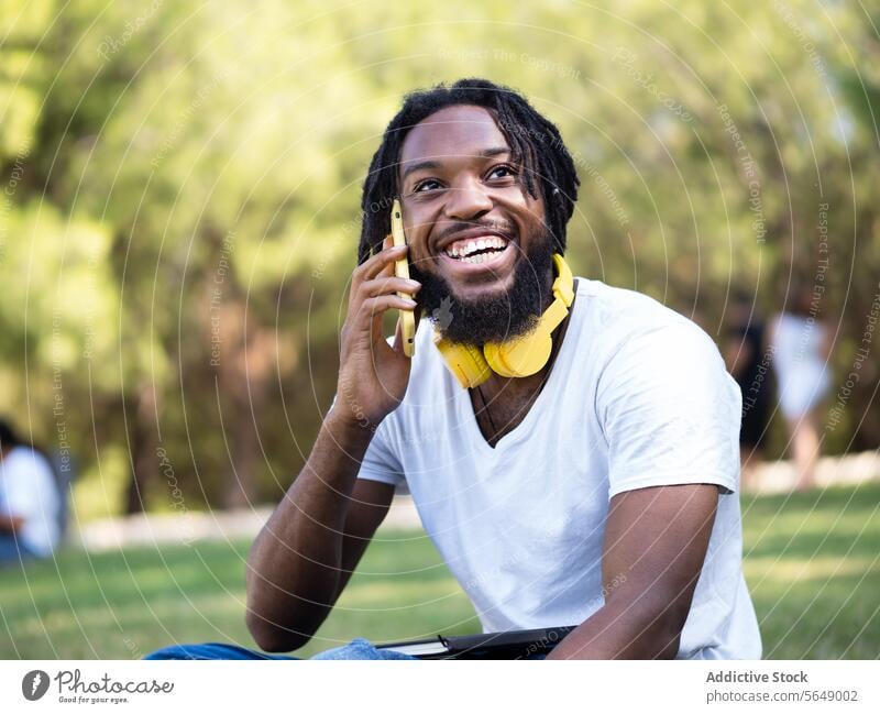 Glücklicher schwarzer Mann führt Telefonat im Park Hipster Kopfhörer benutzend Smartphone Telefonanruf sprechen reden Sommer Lächeln genießen heiter männlich