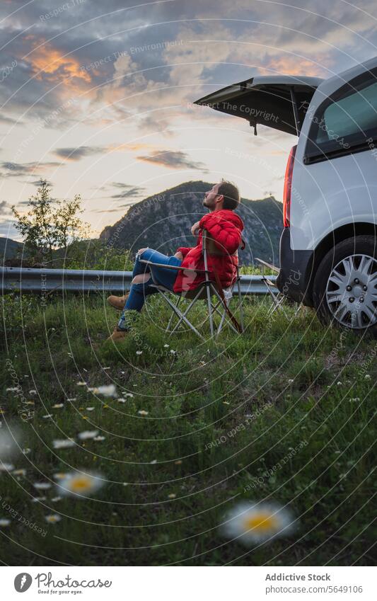 Mann entspannt sich auf einem Stuhl in der Nähe eines Autos in der Abenddämmerung Tourist sich[Akk] entspannen PKW Berge u. Gebirge grasbewachsen Autoreise Feld