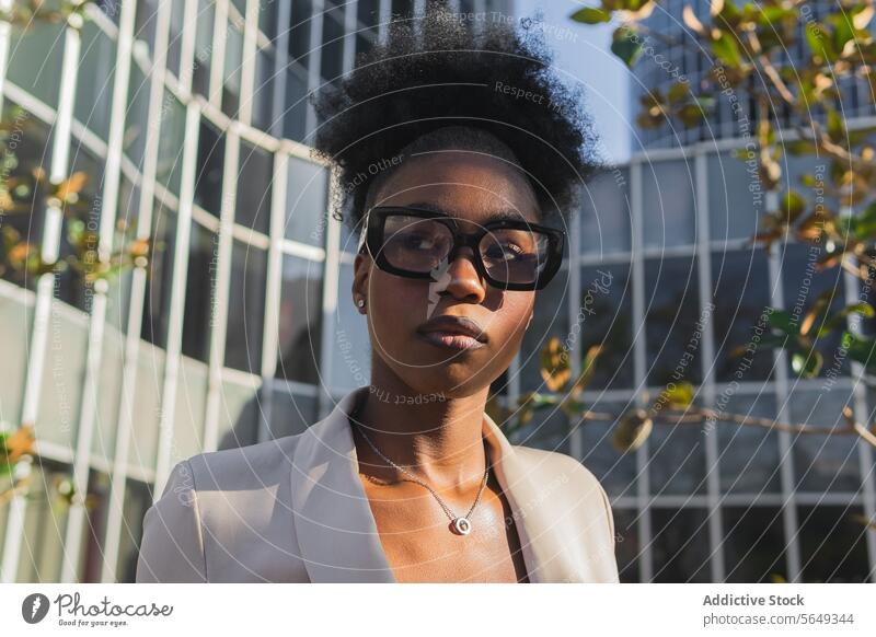 Seriöse schwarze Geschäftsfrau mit Brille und Blazer, die wegschaut, während sie an einem Gebäude in der Stadt steht Unternehmer lockig Behaarung Brötchen