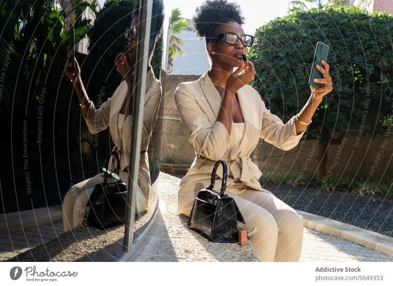 Schwarze Unternehmerin in eleganter Freizeitkleidung trägt Lippenstift auf und benutzt ihr Smartphone als Spiegel, während sie im Freien sitzt professionell