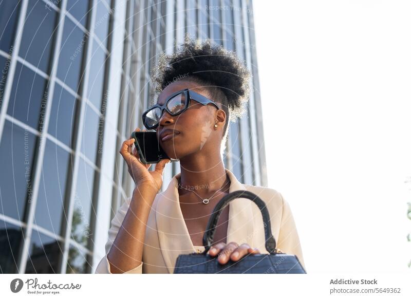 Niedriger Winkel des jungen schwarzen weiblichen Managers, der mit einem Mobiltelefon spricht und an einem Gebäude sitzt Geschäftsfrau Smartphone Geldbörse