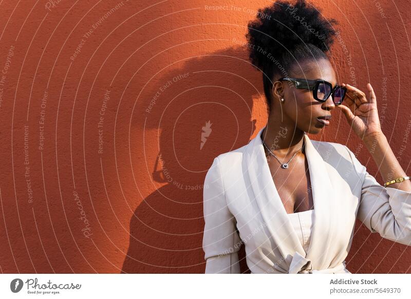 Stilvolle schwarze Geschäftsfrau, die eine Brille gegen eine Wand mit Schatten einstellt Unternehmer rot modisch Behaarung lockig stylisch Textfreiraum Beruf