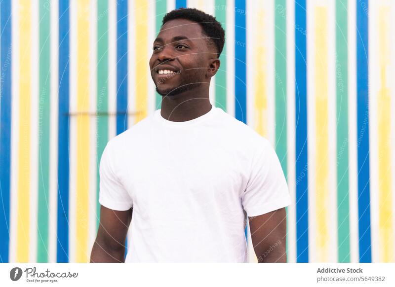 Heiterer schwarzer Mann vor gestreifter Wand Streifen Großstadt Straße urban farbenfroh Geometrie Muster tagsüber Zahnfarbenes Lächeln heiter Glück positiv
