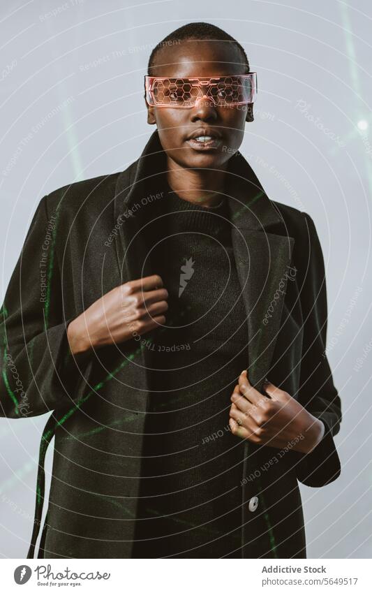 Porträt einer selbstbewussten jungen, mutigen afroamerikanischen Frau mit einer intelligenten, futuristischen VR-Brille und einem langen schwarzen Trenchcoat, die in die Kamera blickt, vor einem mit Neonlicht beleuchteten Hintergrund