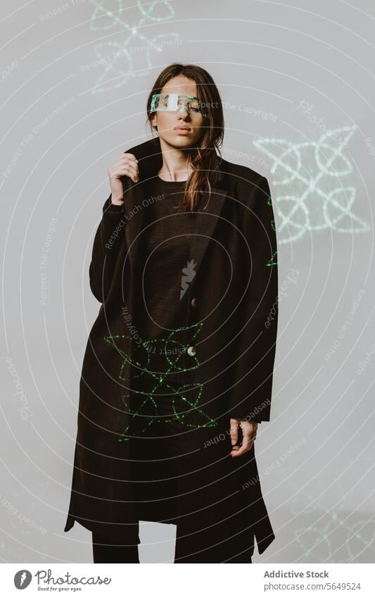 Porträt einer jungen Frau mit intelligenter futuristischer VR-Brille und langem schwarzen Trenchcoat, die mit geschlossenen Augen vor einem beleuchteten Hintergrund steht