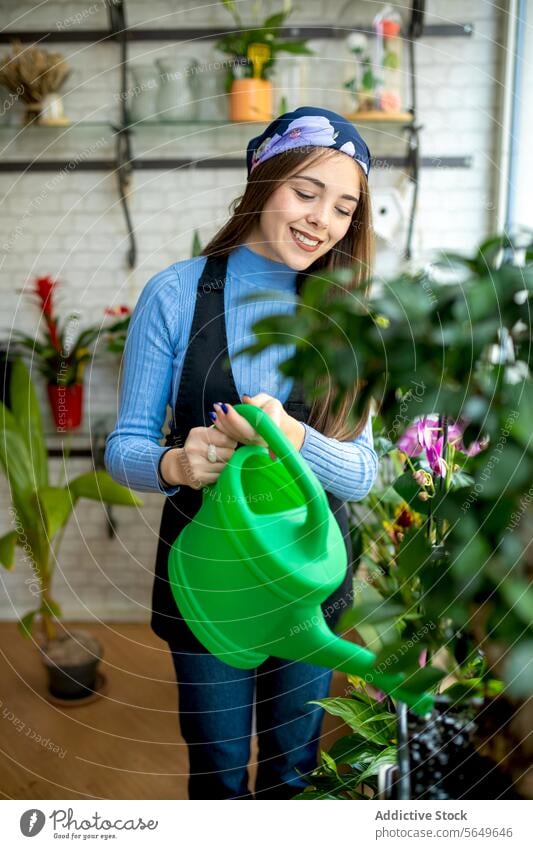 Lächelnde Blumenhändlerin beim Gießen von Topfpflanzen Frau Blumenladen Gießkanne Wasser eingießen eingetopft Pflanze Pflege Arbeitstag jung Schürze