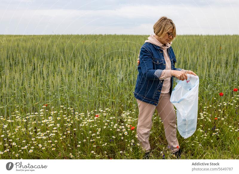 Erwachsene Frau steht mit einem Müllsack und sammelt Plastikabfälle für das Recycling Kunststoff Abfall Wiese Wildblumen Umwelt Erhaltung Aktivität