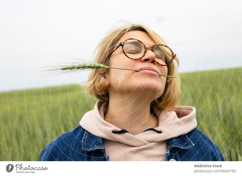 Erwachsene Frau mit einem Weizenhalm im Mund in einem Weizenfeld stehend Stengel Zähne Lächeln Brille Jeansjacke Kapuzenpulli Erholung Genuss Feld Natur einfach