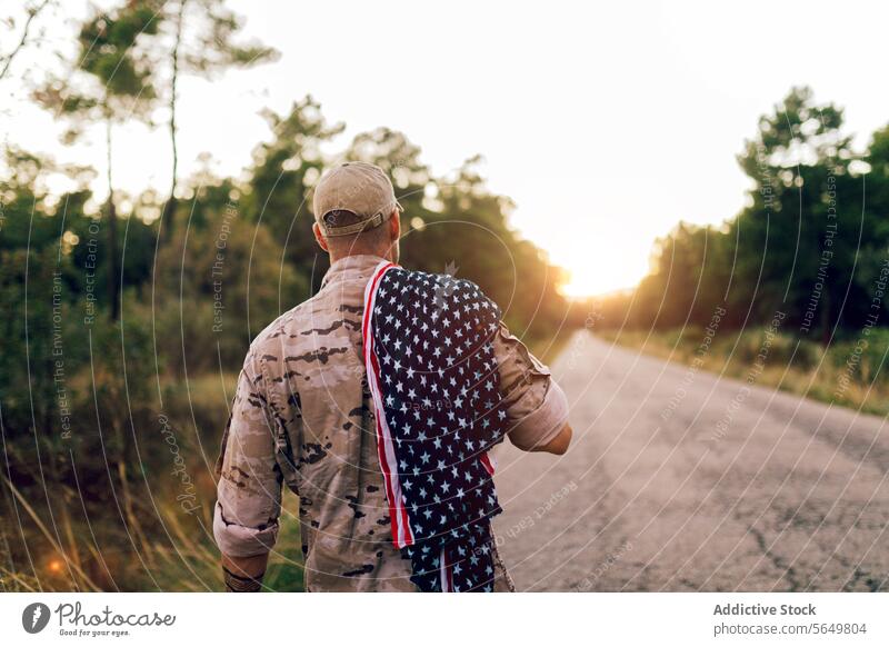Rückenansicht eines unkenntlichen reifen Kommandos auf der Straße mit amerikanischer Flagge auf der Schulter unter dem Himmel bei Sonnenuntergang Mann Soldat