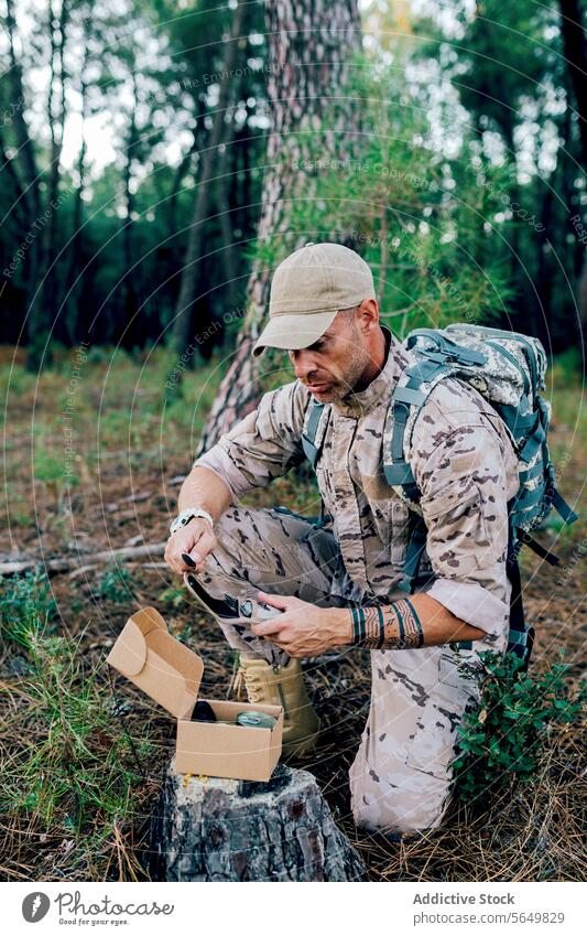 Militärsoldat mit Rucksack und Bestecktasche kniend vor einer Kiste auf einem Baum Mann Kommando Kasten Beutel Tarnung Wald Stumpf Uniform Soldat zwängen