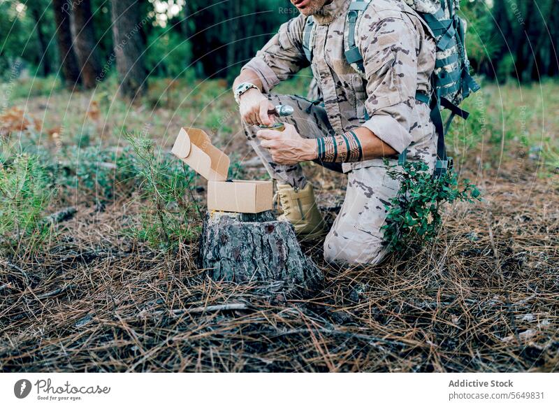 Anonymer Militärsoldat mit Rucksack, der eine Konservendose öffnet, kniend neben einer Kiste auf einem Baum Mann Kommando Besteck Kasten Tarnung Wald Uniform