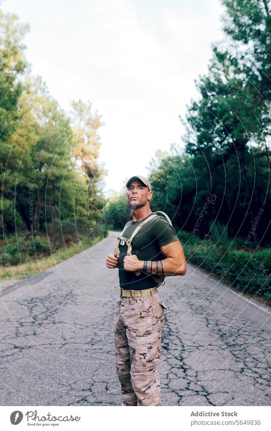 Armee-Mann in Uniform mit Rucksack und Mütze steht auf der Straße gegen Bäume im Wald Soldat selbstbewusst Baum Tarnung keuchen grün Wegsehen Militär Krieger