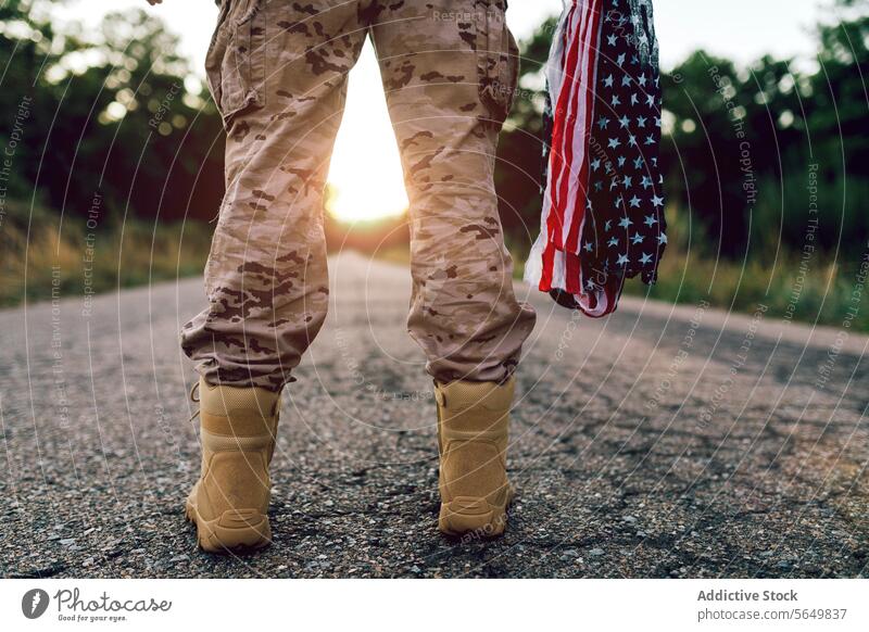 Crop-Beine eines anonymen Kriegers mit Fahne, der bei Sonnenuntergang auf der Straße steht Mann Kommando Amerikaner Tarnung Stiefel Uniform Frontlinie
