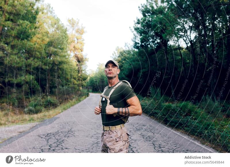 Armee-Mann in Uniform mit Rucksack und Mütze steht auf der Straße gegen Bäume im Wald Soldat selbstbewusst Baum Tarnung keuchen grün Wegsehen Militär Krieger