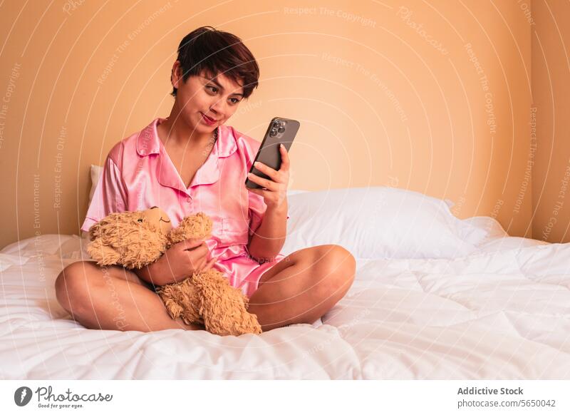 Lächelnde junge Frau mit weichem Teddybär beim Surfen auf dem Smartphone im Schlafzimmer benutzend Browsen zu Hause soziale Netzwerke Glück Telefon Internet