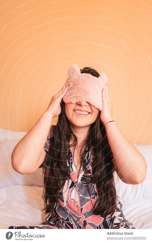 Lächelnde Frau mit Schlafmaske auf dem Bett sitzend Pyjama Augen abdecken Nachtwäsche Mundschutz Komfort gemütlich Schlafzimmer jung heiter Freude wach Glück