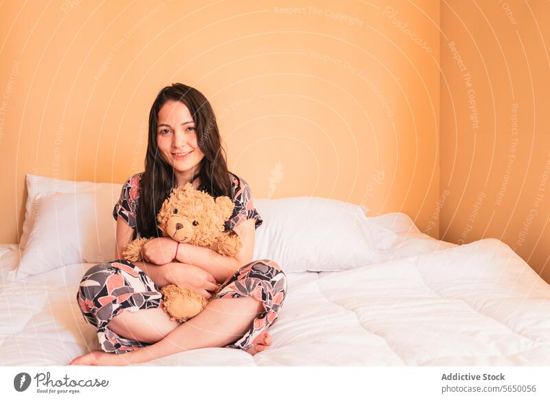 Junge Frau sitzt mit Teddybär auf dem Bett Bär Spielzeug Umarmung Lächeln Plüsch gemütlich Schlafzimmer jung Komfort Glück Pyjama Nachtwäsche