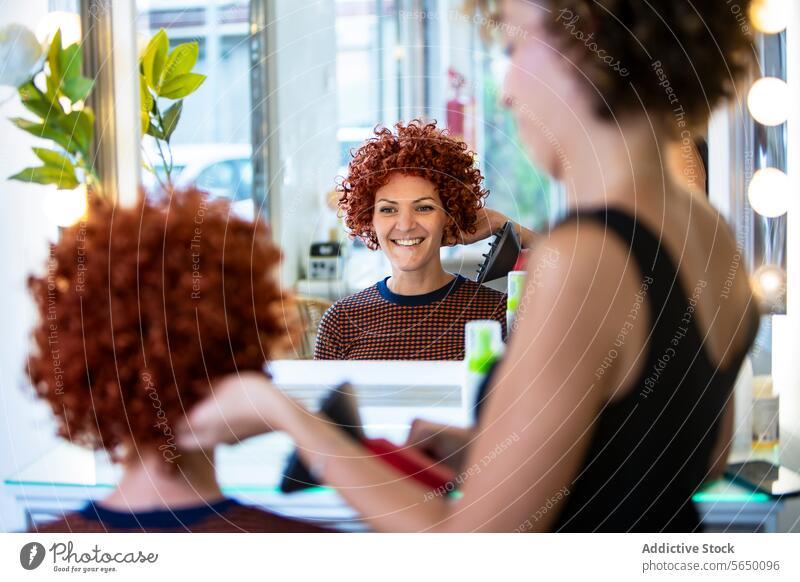 Lächelnde Frau, die ihr lockiges Haar im Spiegel eines Salons stylt krause Haare Haarstyling Haarglätter Friseursalon heiter stylen modern