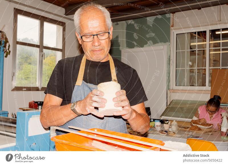 Älterer Handwerker bei der Arbeit in einer Töpferwerkstatt Kunsthandwerker Kunstgewerbler Frau Ton Vase Schaber zerkratzen Keramik Schürze Zusammensein