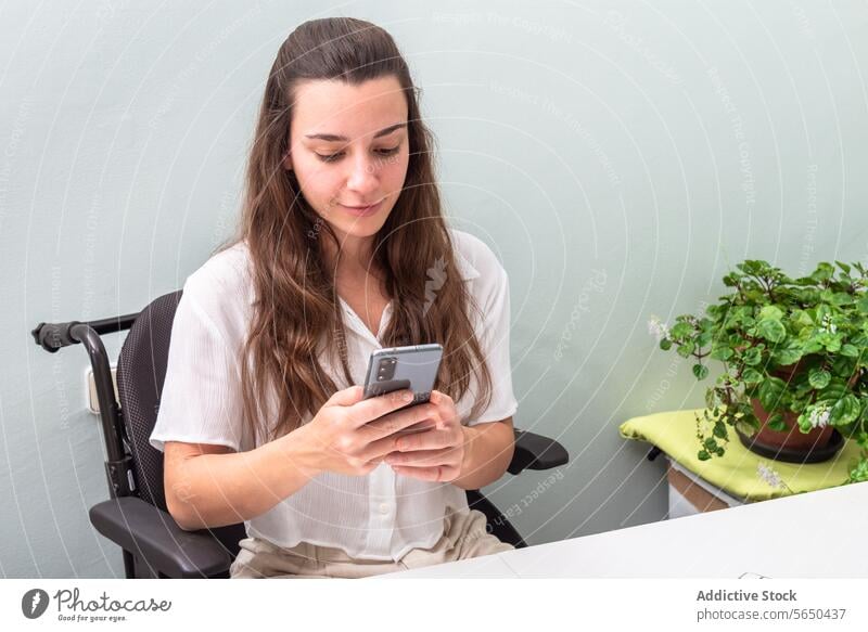 Berufstätige Frau im Rollstuhl beim Telefonieren an einem modernen Arbeitsplatz Smartphone Browsen Schreibtisch Büro Pflanze sitzend jung lässig professionell