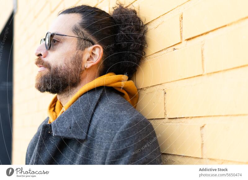 Moderner Mann mit Sonnenbrille, der sich gegen eine gelbe Backsteinmauer lehnt Stil modern Wand Baustein grau Mantel Schal nachdenklich Tag sonnig Hipster urban