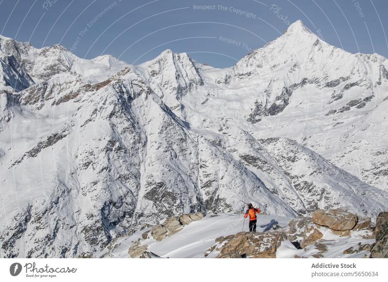 Ganzer Körper eines unerkennbaren Rucksacktouristen in warmer Kleidung, der auf einem Felsen vor verschneiten Bergen steht, während eines Urlaubs in Zermatt