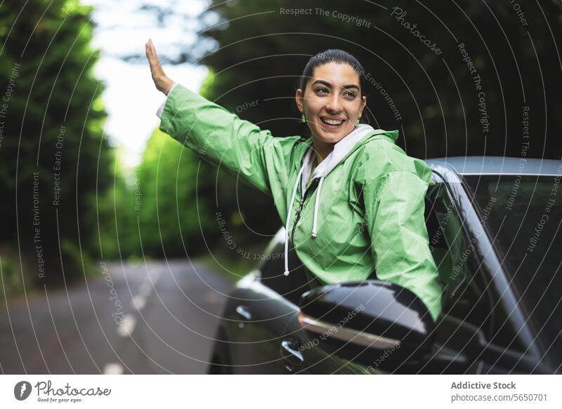 Glückliche junge Frau mit ausgestreckten Armen, die wegschaut und sich aus dem Autofenster lehnt, während sie einen Road Trip im Wald genießt sorgenfrei