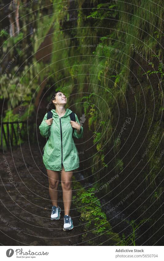 Lächelnde junge Frau in grünem Regenmantel blickt in voller Länge nach oben, während sie im Urlaub an einer mit Moos bewachsenen Klippe wandert wandern