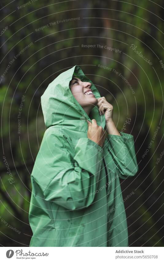 Lächelnde junge Frau in grünem Regenmantel mit Kapuzenpulli, die beim Wandern an einer mit Moos bewachsenen Klippe nach oben schaut wandern aufschauend Nachlauf