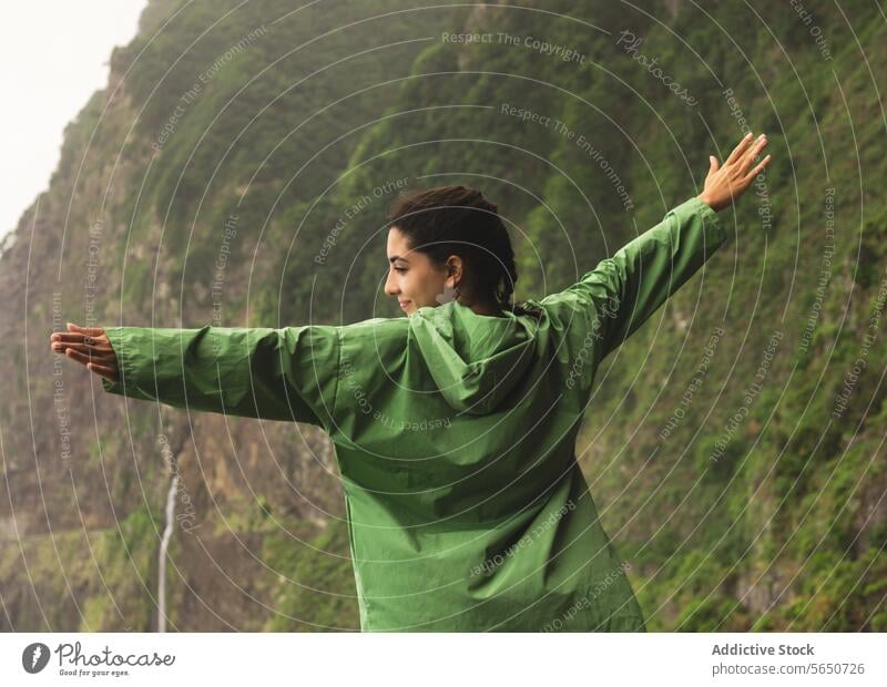 Rückenansicht einer unbekümmerten jungen Frau in grünem Regenmantel, die mit ausgestreckten Armen auf einer Betonmauer vor Bergen steht sorgenfrei Barriere
