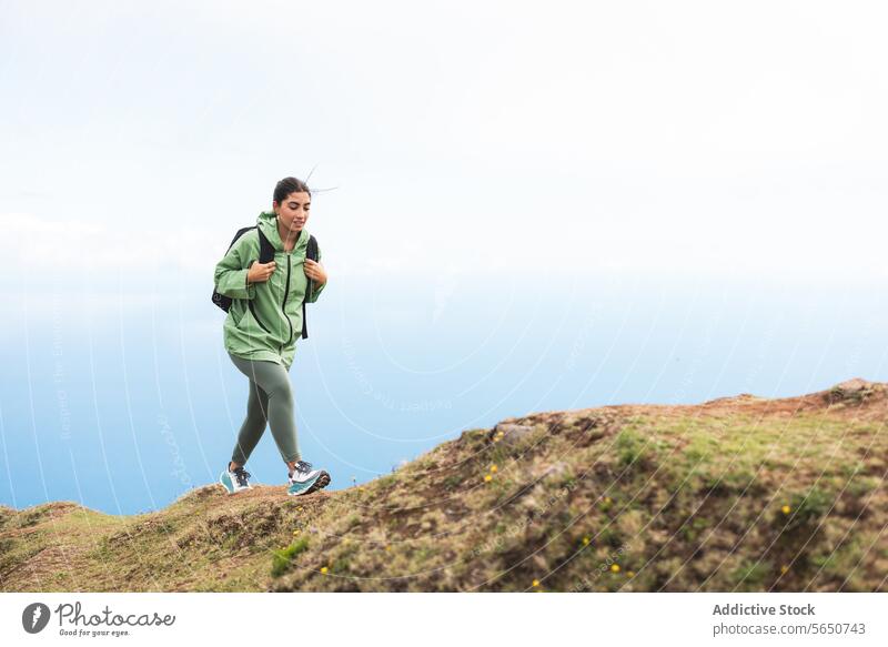 Ganzkörper-Seitenansicht einer jungen Wanderin in grünem Regenmantel mit Rucksack, die sich während eines Wochenendurlaubs auf einem Berggipfel gegen den Himmel bewegt