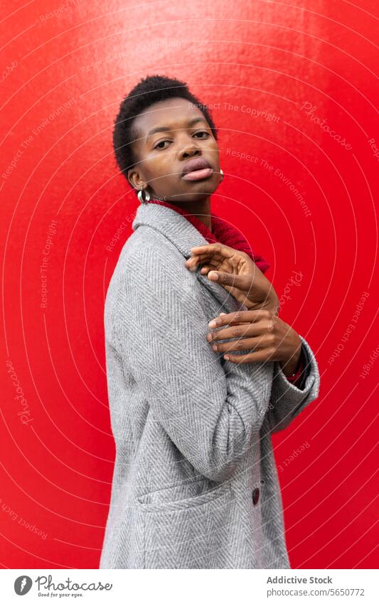 Seitenansicht einer emotionslosen afroamerikanischen Frau mit kurzen Haaren in einer warmen Jacke und Ohrringen, die in die Kamera schaut und auf einem roten Hintergrund steht