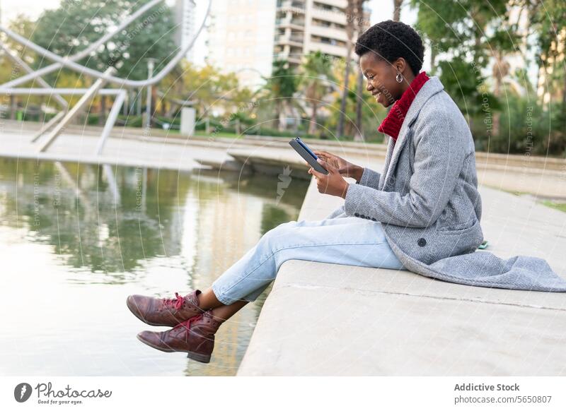 Seitenansicht einer schwarzen Frau, die ein elektronisches Buch im Park liest benutzend digitales Buch e-Buch lesen Großstadt Teich Lächeln positiv Glück