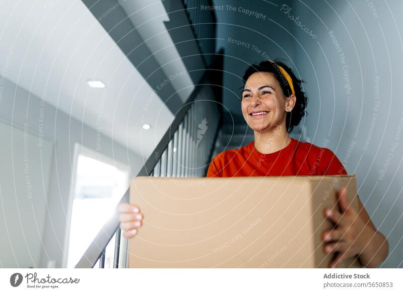 Lächelnde Frau mit Kiste auf der Treppe in neuer Wohnung Karton Kasten führen verlegen einziehen heimwärts Treppenhaus heiter Zugehörigkeit Appartement