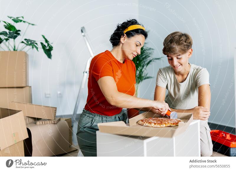 Glückliche Mutter beim Pizzaschneiden mit Cutter und Sohn schaut mit Freude zu geschnitten appetitlich auspacken Zugehörigkeit einziehen neu flach Appartement