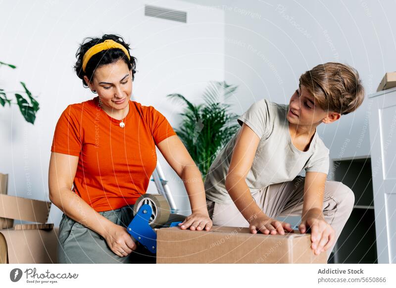 Fokussierte Mutter in roter Top-Verpackungsbox mit Klebebandabroller mit Sohn Kasten Hilfsbereitschaft Spender Appartement neu Paket Teenager sich[Akk] bewegen