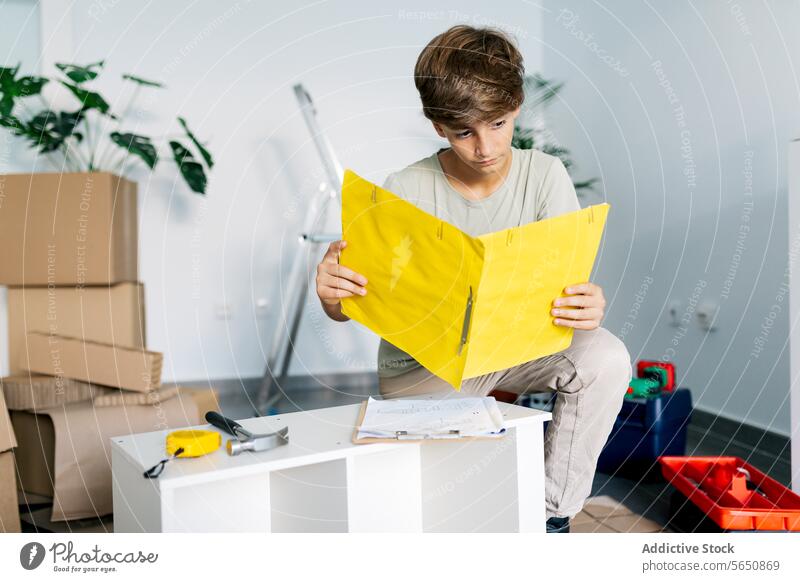 Konzentrierter Junge kniend und Notizen auf gelbem Klemmbrett lesend Belehrung versammeln Möbel Hinweis Zwischenablage prüfen verlegen Teenager Fokus