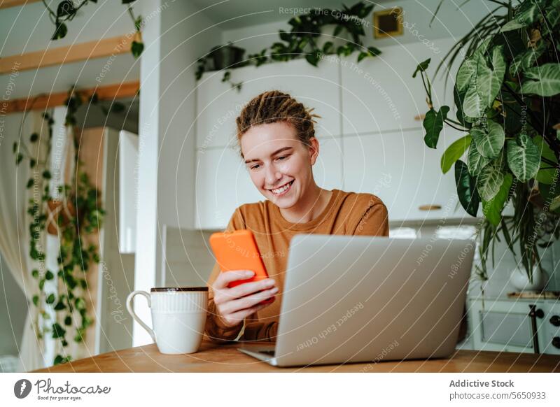 Lächelnde junge Frau mit Smartphone am Küchentisch mit Laptop bei Tageslicht Glück Textnachricht benutzend online zu Hause sitzen Rastalocken Browsen Surfen
