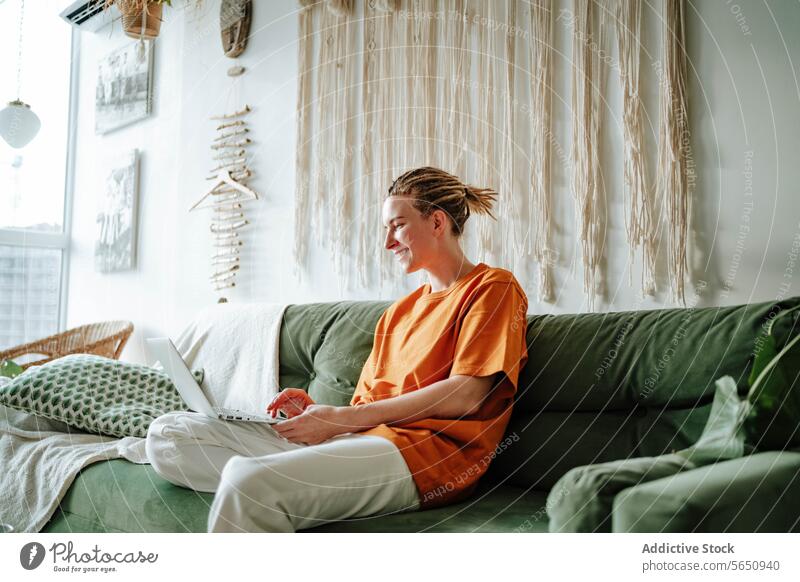 Junge, lächelnde Frau mit Laptop auf dem Sofa zu Hause in der Nähe des Fensters freiberuflich Lächeln benutzend Arbeit Projekt abgelegen Netbook pc Glück