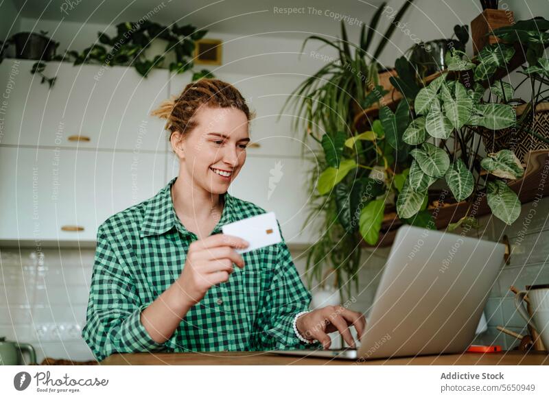 Junge Frau sitzt mit Kreditkarte in der Nähe von Laptop am Tisch im Tageslicht zu Hause Kauf online Lächeln Werkstatt Glück froh jung sitzen positiv Gerät