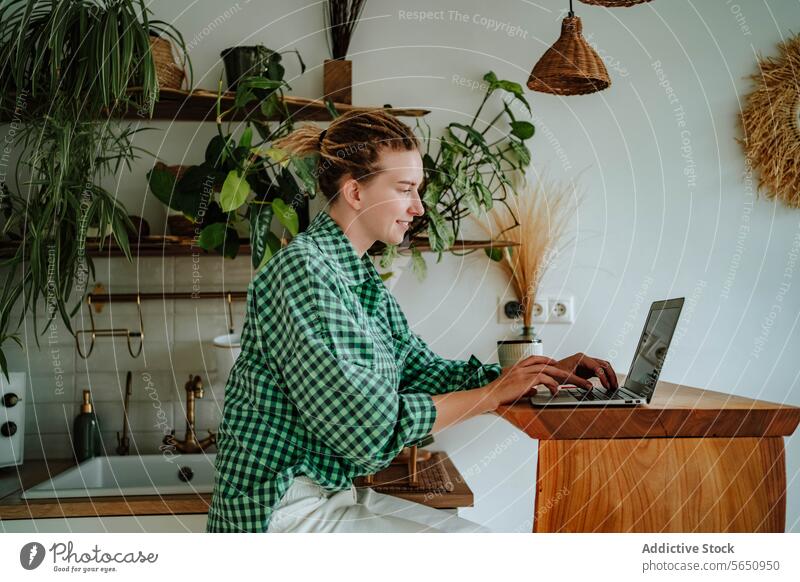 Glückliche junge Frau arbeitet am Laptop am Tisch in der dekorierten Küche zu Hause freiberuflich Arbeit abgelegen Inbetriebnahme online sitzen kariertes Hemd