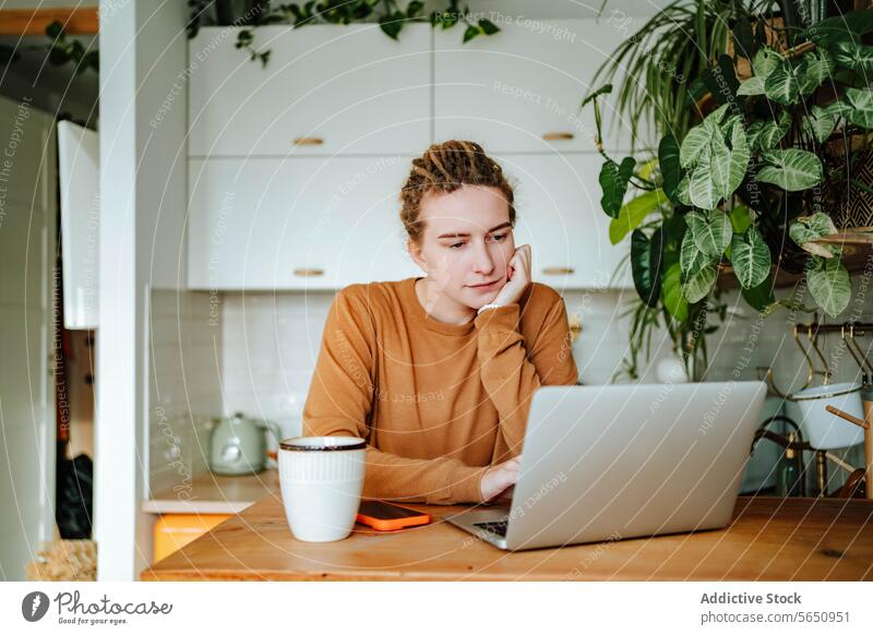Junge Frau arbeitet am Laptop am Küchentisch mit Smartphone Kaffeetasse im Tageslicht nachdenklich sich auf die Hand lehnen Kabinett Rezept Tisch zu Hause jung