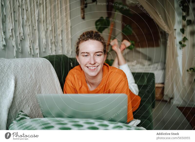 Lächelnde Frau arbeitet am Laptop, während sie zu Hause auf dem Sofa liegt benutzend online abgelegen Arbeit freiberuflich Browsen Internet Glück