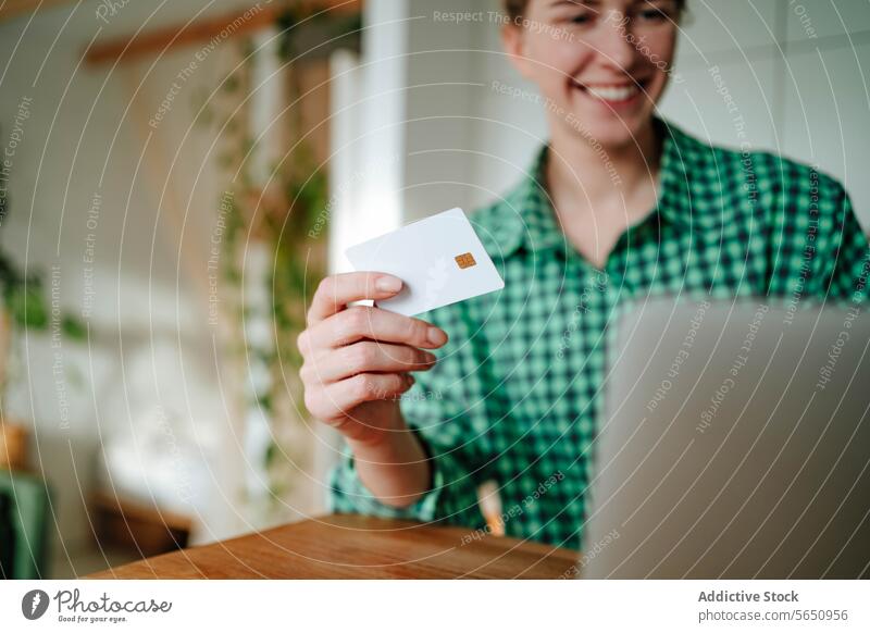 Unscharfe glückliche junge Frau sitzt mit Kreditkarte in der Nähe von Laptop am Tisch im Tageslicht Kauf online Lächeln Werkstatt Glück froh zu Hause sitzen