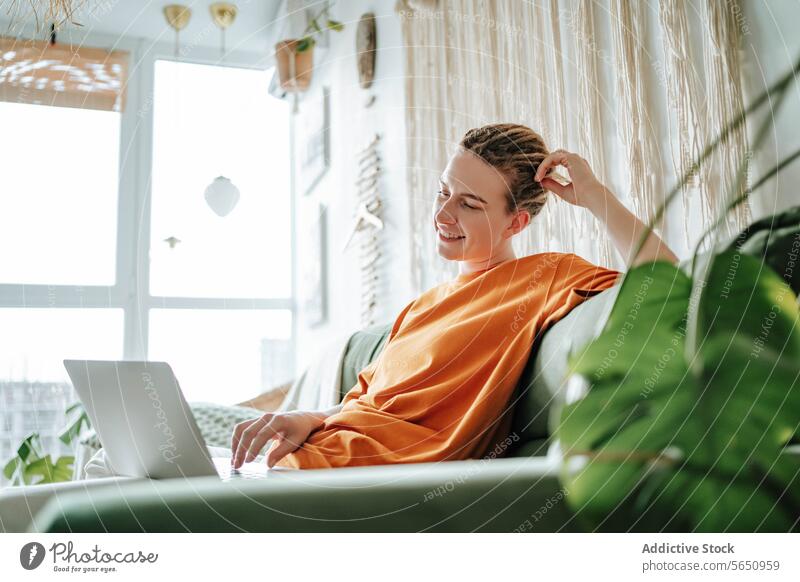 Junge, lächelnde Frau mit Laptop auf dem Sofa zu Hause in der Nähe der Anlage freiberuflich Lächeln benutzend Arbeit Projekt abgelegen Netbook pc Glück Komfort