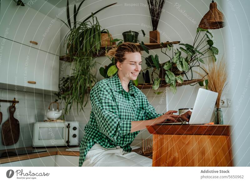 Glückliche junge Frau arbeitet am Laptop am Tisch in der dekorierten Küche zu Hause freiberuflich Arbeit abgelegen Inbetriebnahme online sitzen kariertes Hemd