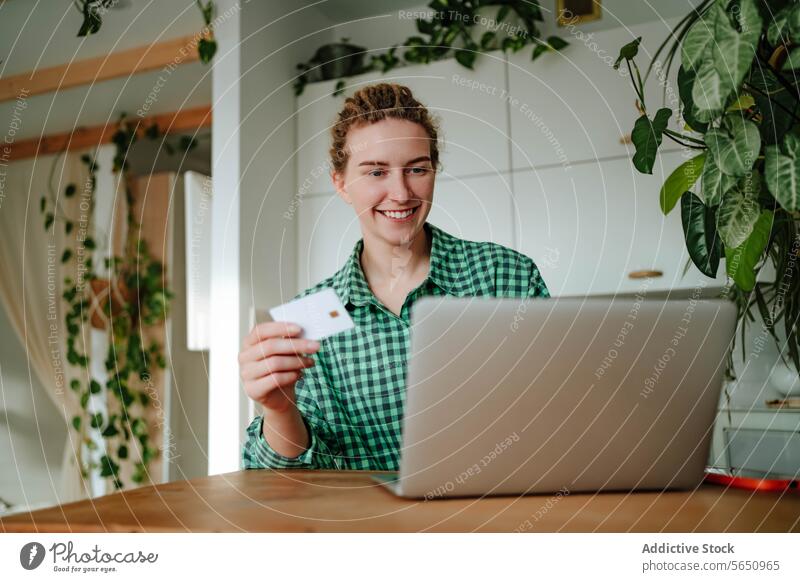 Junge Frau sitzt mit Kreditkarte neben Laptop an einem Tisch im Tageslicht Kauf online Lächeln Werkstatt Glück froh zu Hause jung sitzen positiv Gerät Apparatur